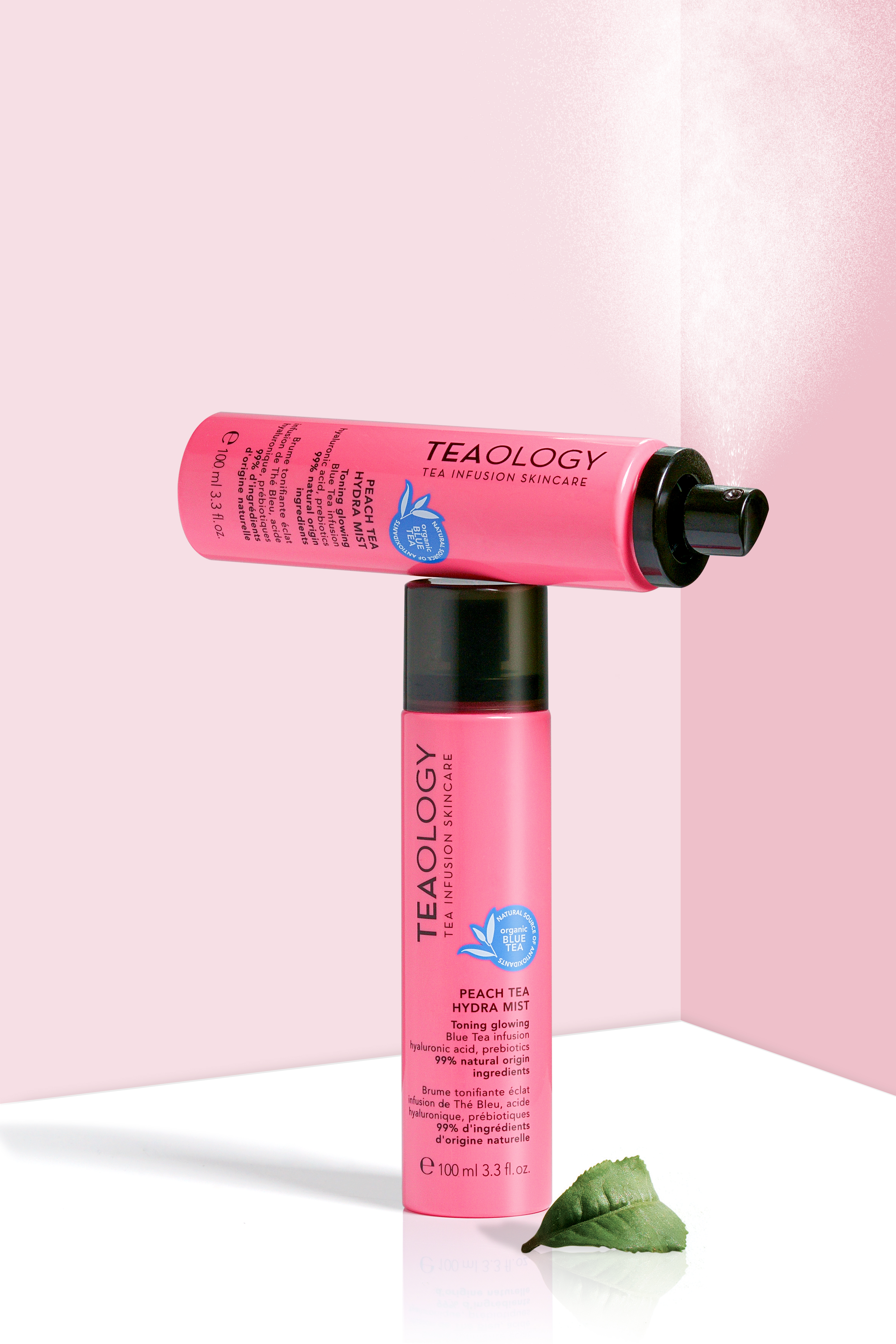 Zwei rosa Flaschen Teaology Peach Tea Hydra Mist auf rosa Hintergrund, eine stehend, eine liegend, mit grünem Blatt.