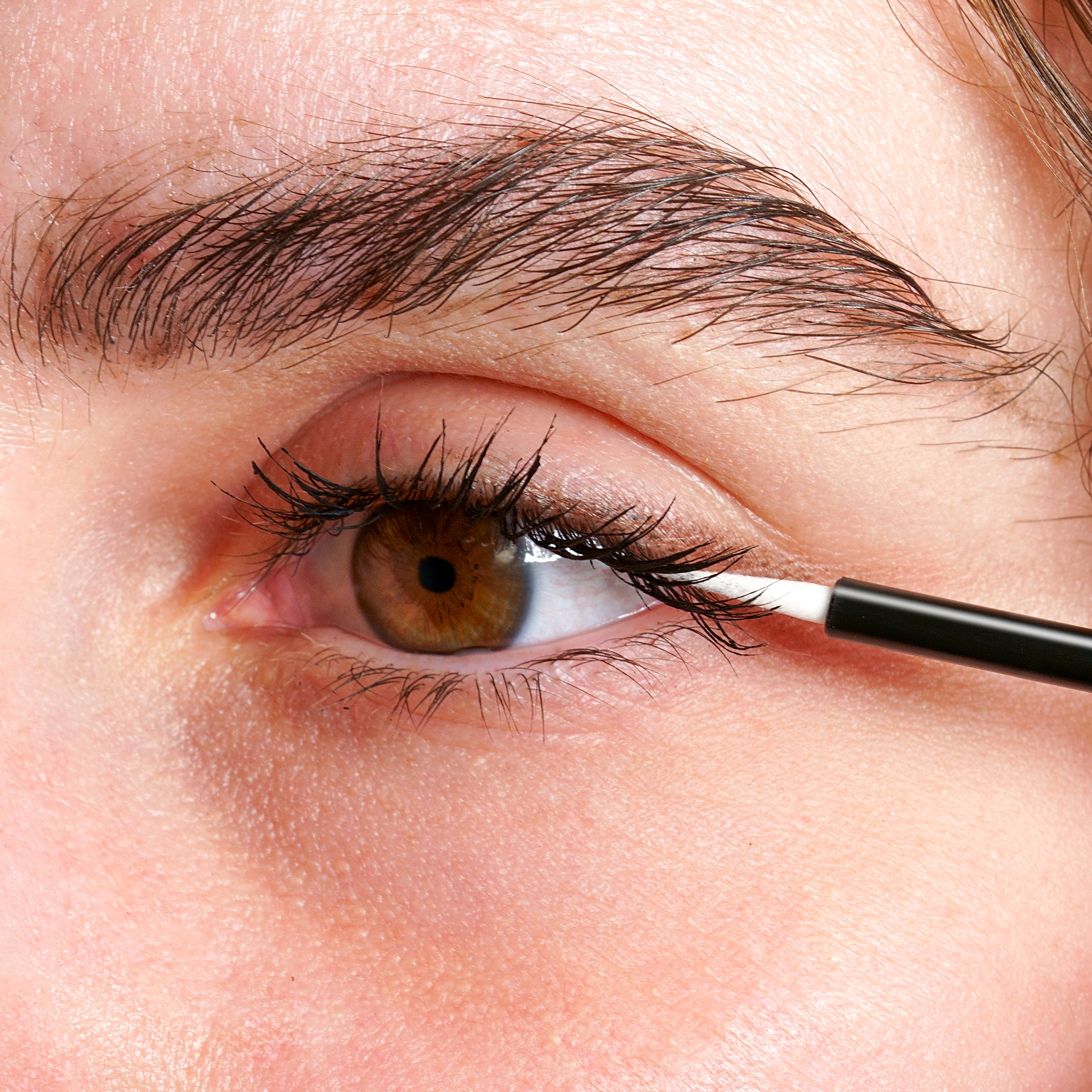 Nahaufnahme eines Auges mit brauner Iris, auf das ein Pinsel Wimpernserum aufträgt.