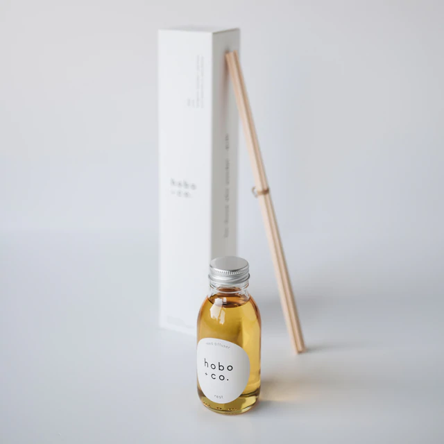 Kleine Flasche mit Duftöl von hobo + co. und Holzstäbchen vor einer weißen Verpackung auf einem weißen Hintergrund.